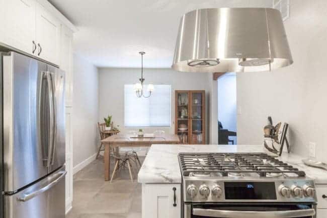 Modern kitchen with a stainless steel refrigerator, gas stove, and marble countertop in the foreground; perfect inspiration for your next kitchen renovation. Dining area features wooden table, white chairs, and a large window in the background.