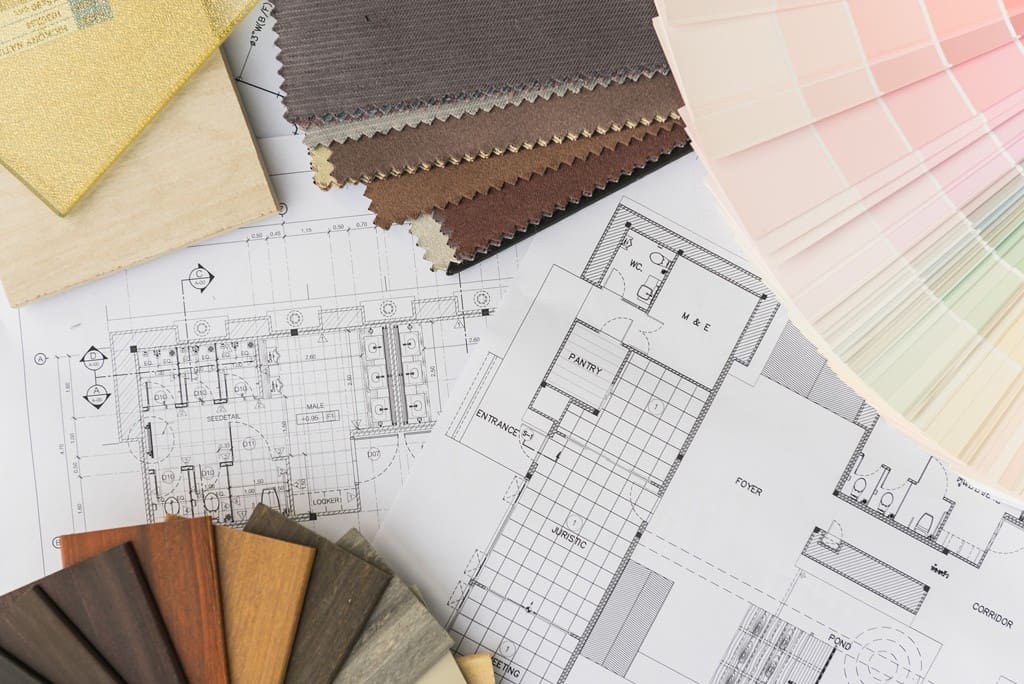 Interior design planning materials laid out, including architectural blueprints, fabric swatches, wood samples, and a color palette fan—each playing a key role in visualizing style and estimating project cost—all arranged on a flat surface.