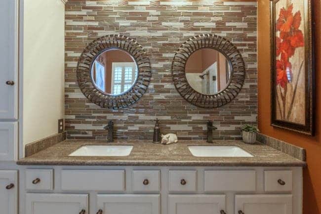 Experience an award-winning transformation in this Ocotillo Lakes bathroom vanity, featuring two sinks, round decorative mirrors, wall-mounted faucets, a mosaic tile backsplash, white cabinets, and a framed red floral painting.