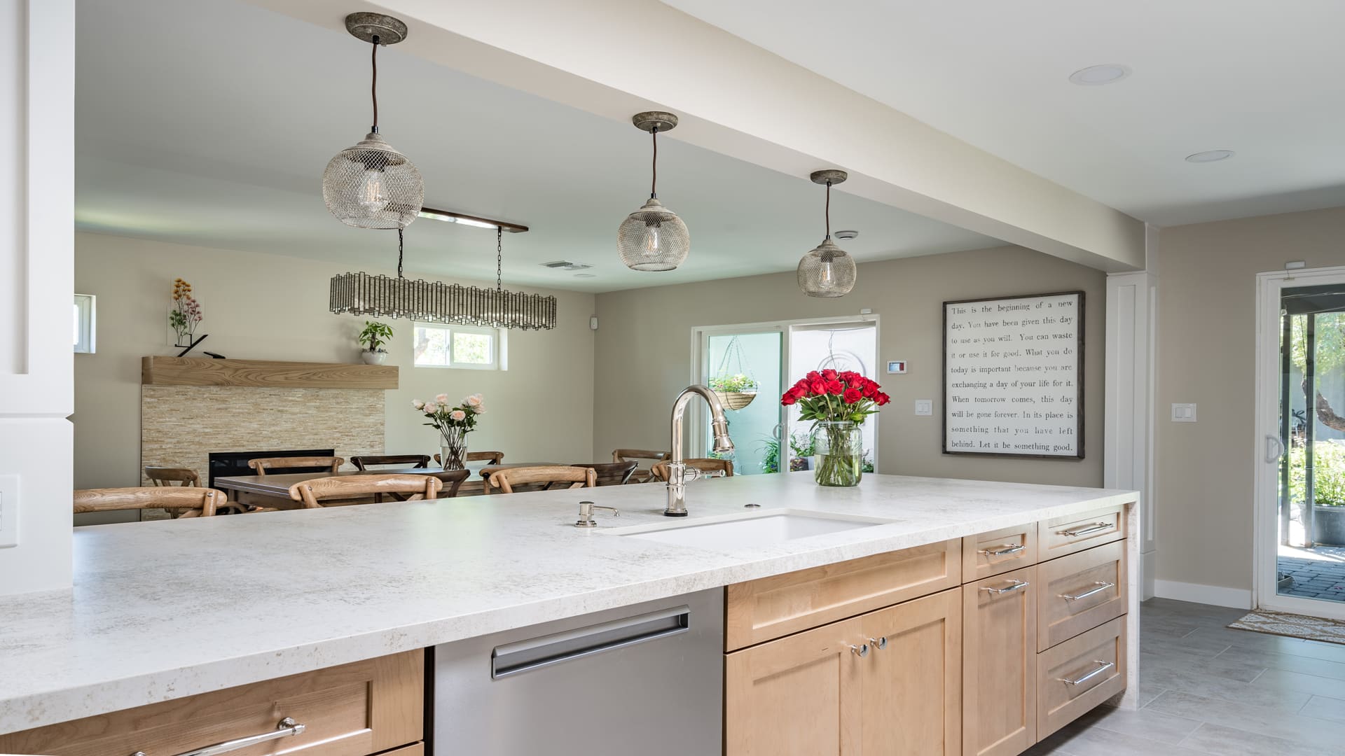 Modern kitchen with light wood cabinets, a white countertop, stainless steel appliances, pendant lights, a vase of red flowers, and an open view to a dining area with a large wall art piece and natural light.