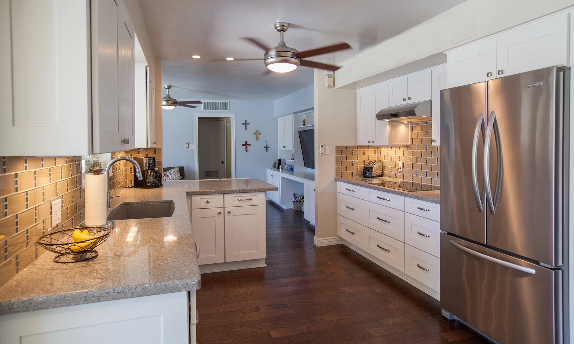 Modern open concept galley kitchen with white cabinets, stainless steel appliances, gray countertops, and a tile backsplash. Dark wood floors and ceiling fans are visible, with fruit in a bowl and decor on the walls in the background.