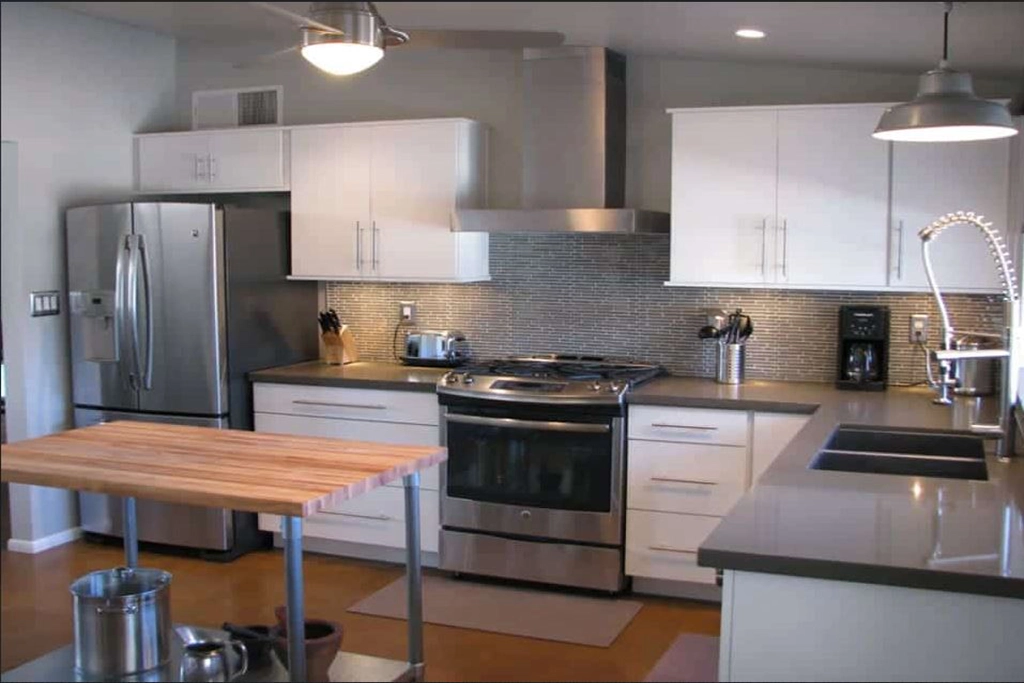 Modern kitchen in Phoenix with stainless steel appliances, white cabinets, gray countertops, a tile backsplash, a Mid Century Modern center island with a wooden top, and gooseneck faucet above a double sink. Coffee maker and utensils on the counter.