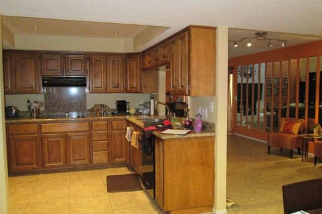 A kitchen with wooden cabinets, granite countertops, and stainless steel appliances. Adjacent is a living area separated by vertical wooden slats, creating an award-winning redesign made for entertaining in this well-lit, tiled space.