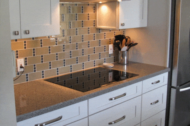 Modern open concept galley kitchen with white cabinets, granite countertop, stainless steel stove and range hood, beige tile backsplash with dark accents, utensil holder, and part of a stainless steel refrigerator visible on the right.