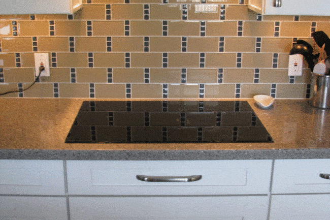Modern open concept galley kitchen with white cabinets, a gray countertop, an electric cooktop, and a stainless steel range hood. The backsplash features a beige and blue geometric tile pattern. Cooking utensils are set in a holder on the right.