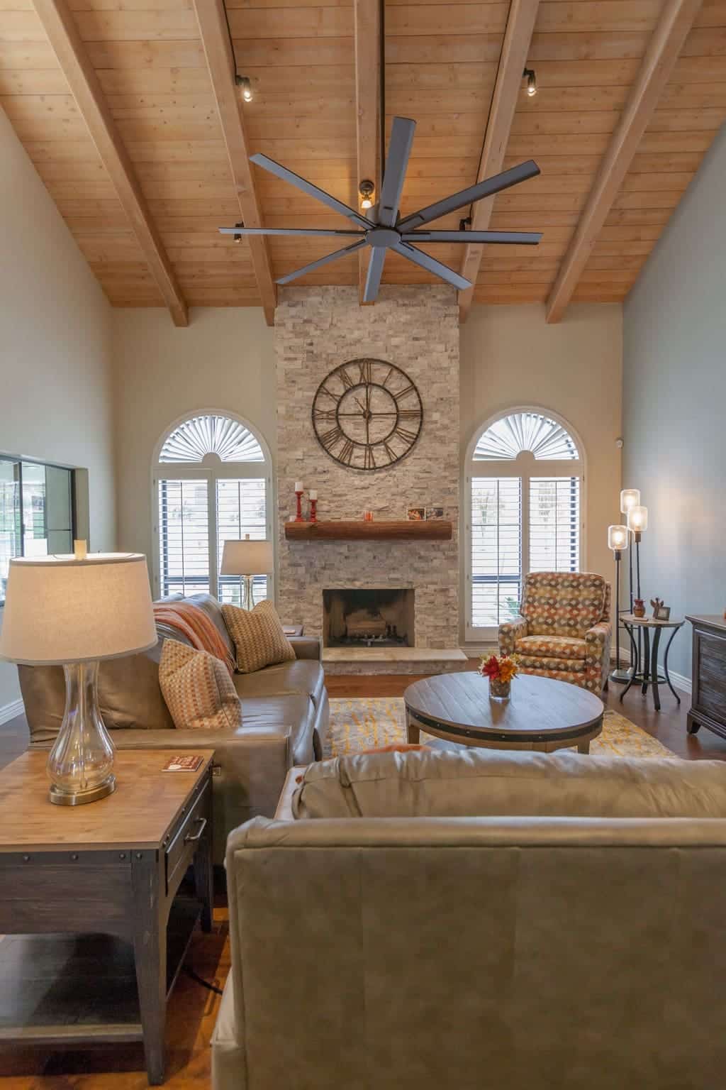 A cozy living room, part of a Contractor of the Year Project, features a vaulted wood ceiling, large fan, stone fireplace, grand clock, sofas, armchair, round coffee table, and arched windows streaming in natural light.