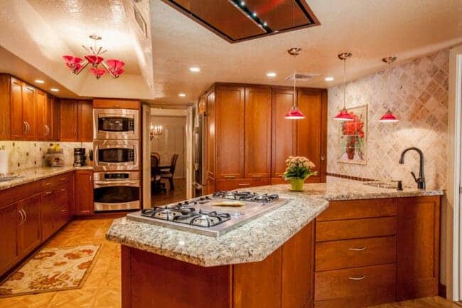 Spacious kitchen with wooden cabinets, granite countertops, a built-in stove on the island, stainless steel appliances, and red pendant lights showcases how the design/build concept works. Dining area visible in the background.