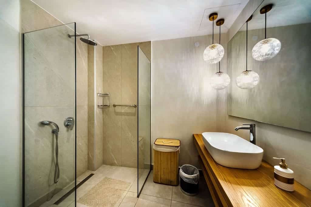 Modern bathroom with a walk-in glass shower highlights one of the 5 REASONS TO CONSIDER A CURBLESS SHOWER, featuring a wooden vanity with vessel sink, round hanging lights above, mirror, waste bins, and neutral-toned tile walls and floor.