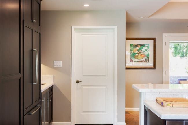 Modern Entertainers Kitchen featuring dark wood cabinets, light countertops, large floor tiles, and neutral walls. A white door is centered at the back, with framed artwork near a glass door leading outside.