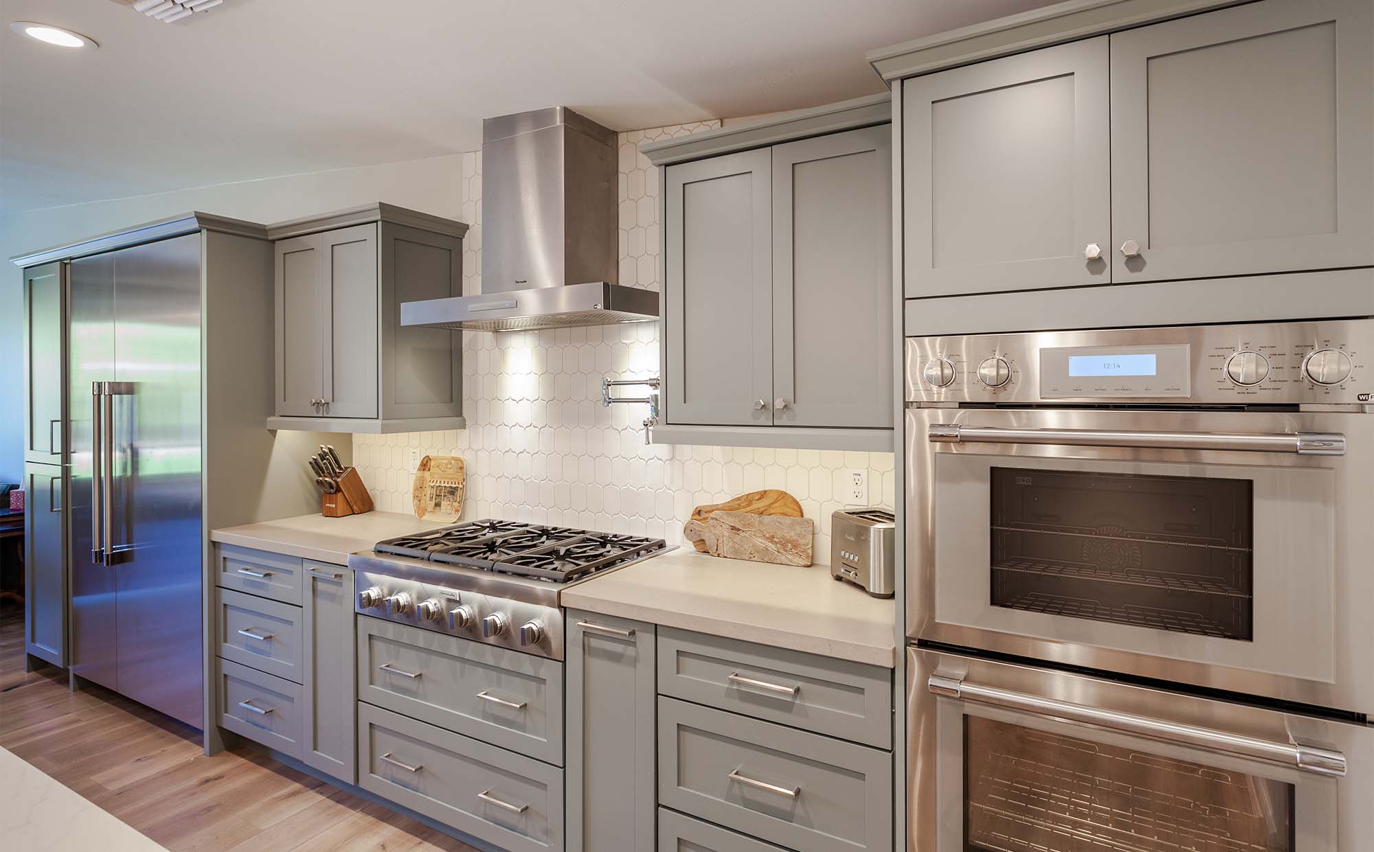Modern kitchen designed for aging in place, with gray cabinets, stainless steel appliances including a double oven and range hood, a large refrigerator, gas stove, white countertops, and light wood flooring. Cutting boards and utensils sit on the counter.