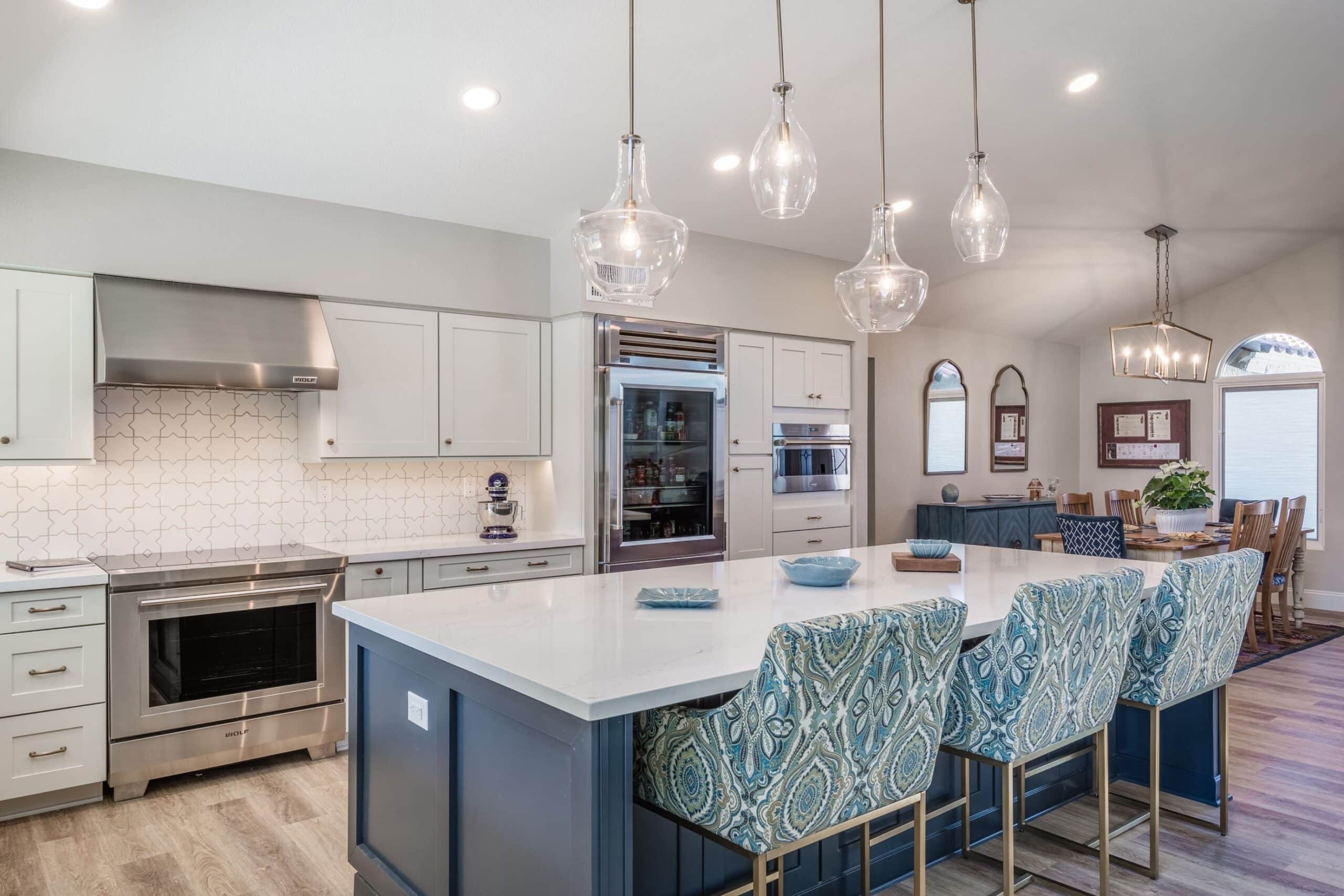 Modern kitchen with a large island, patterned blue barstools, stainless steel appliances, white cabinetry, pendant lights, and an open dining area—part of a stunning Whole Home Remodel.