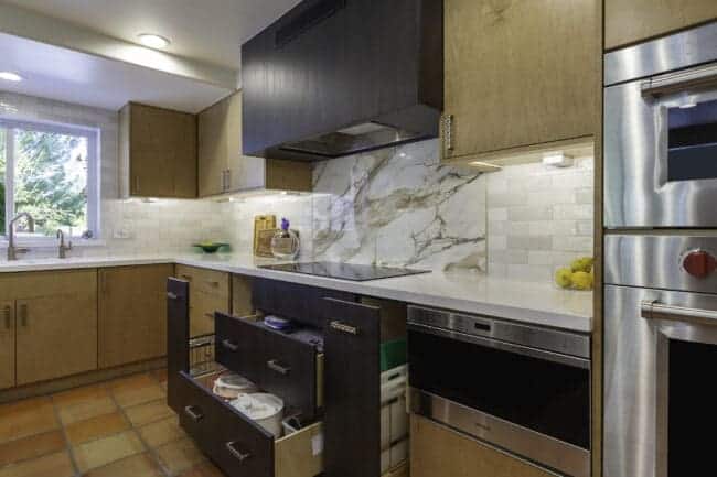Modern custom kitchen with light wood cabinets, marble backsplash, induction cooktop, built-in oven, open drawers showing cookware, and a window letting in natural light. White countertops and tiled floor are also visible.
