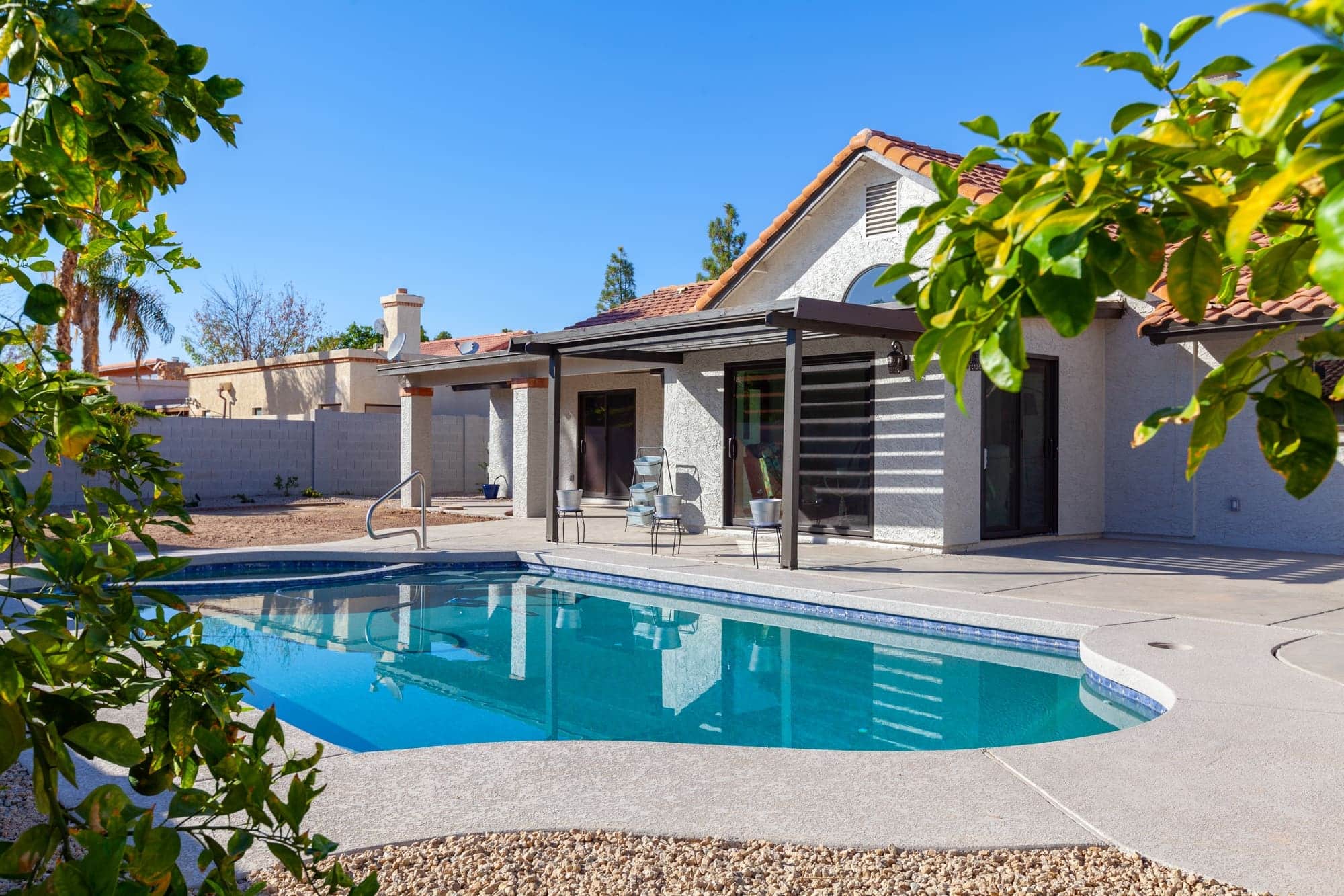 A backyard transformed by a whole home remodel features a curved swimming pool, concrete patio, and covered outdoor seating area, all surrounded by gravel and greenery beside a single-story house with a tiled roof.