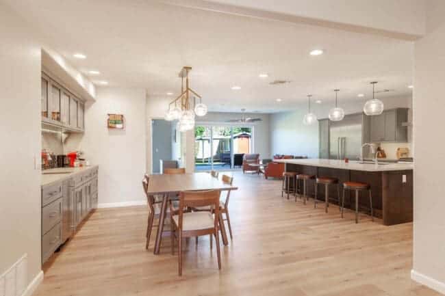 Modern open-concept kitchen and dining area in a mid-century modern home, featuring light wood floors, gray cabinets, a wooden dining table with six chairs, pendant lights, and large windows letting in natural light.