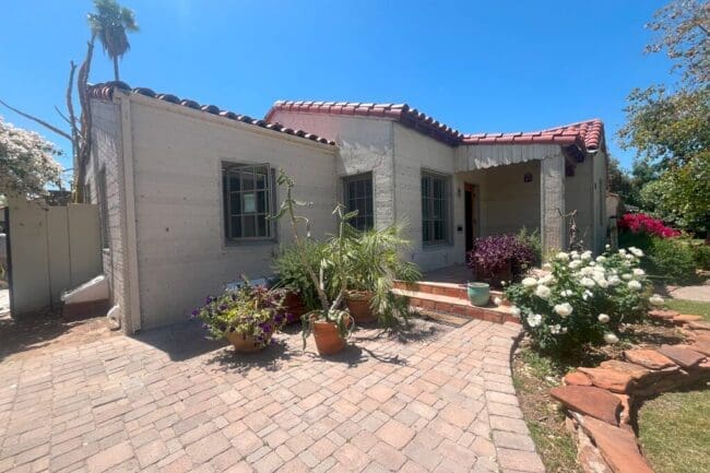 A small house with a red tile roof and light-colored walls, surrounded by potted plants and flowers, sits under a clear blue sky with a paved stone walkway leading to the entrance.