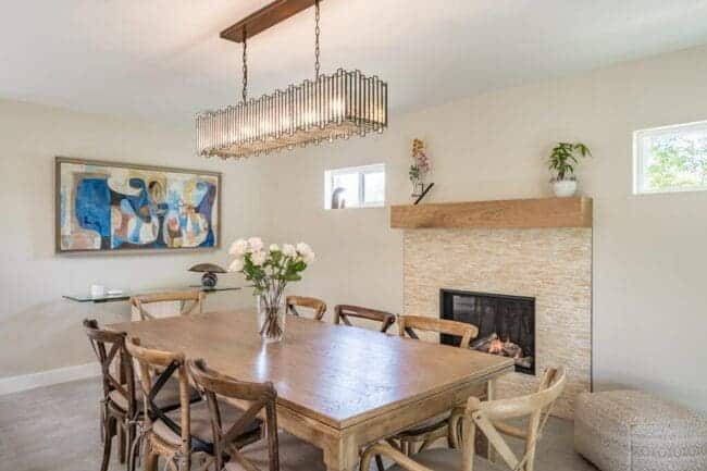 A modern dining room in Paradise Valley featuring a wooden table, eight chairs, a vase of flowers, fireplace, abstract wall art, pendant light, and plants on the mantel. Neutral colors and abundant natural light complete this home remodel.