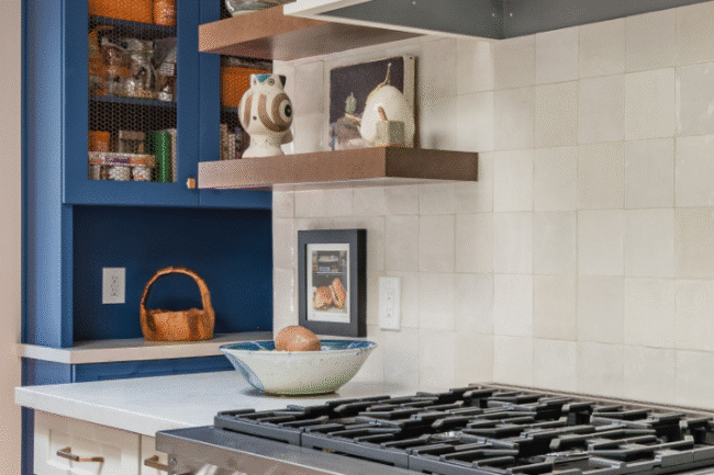 A modern kitchen with a stainless steel gas stove, white cabinets, wooden shelves holding dishes, and a blue cabinet with a mesh door displaying pantry items. A bowl with eggs and a basket are on the counter.
