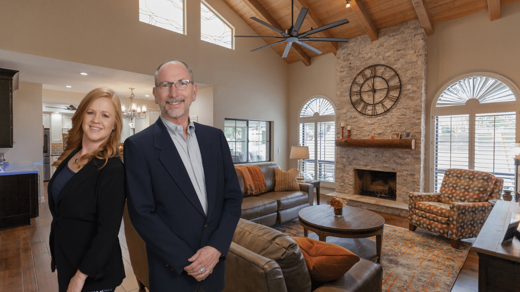 A smiling man and woman in business attire stand in a spacious, modern living room—recently transformed by a leading home remodeling in Phoenix—with vaulted wooden ceilings, a stone fireplace, large windows, and comfortable furniture.
