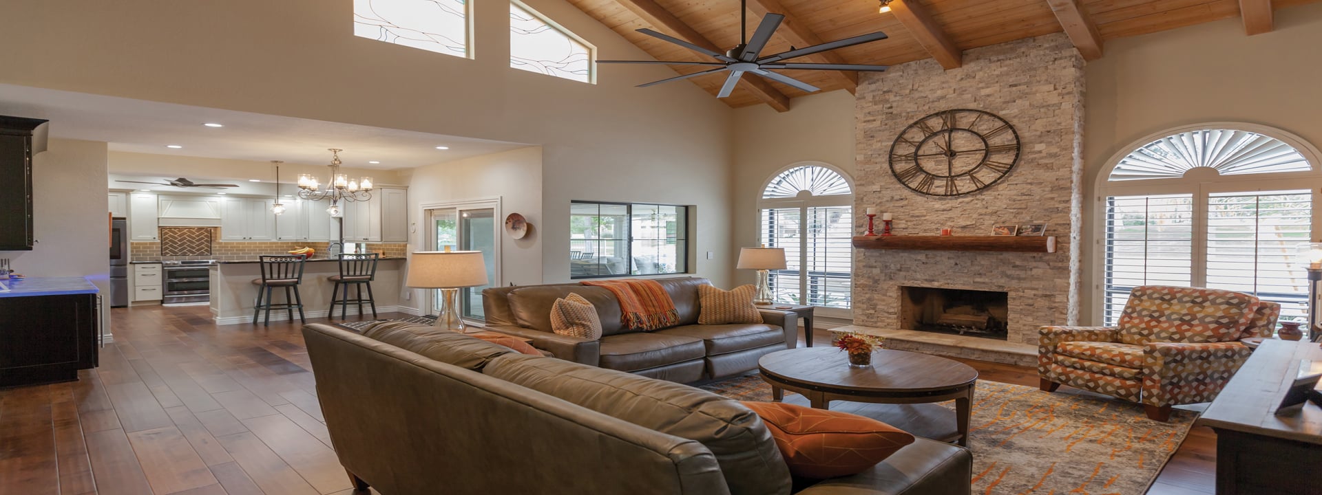 Spacious living room with vaulted wooden ceiling, large ceiling fan, stone fireplace with a large clock, leather sofas, and patterned armchair. Perfect for home remodeling inspiration, this Biltmore-style space boasts sunlight streaming through arched windows.