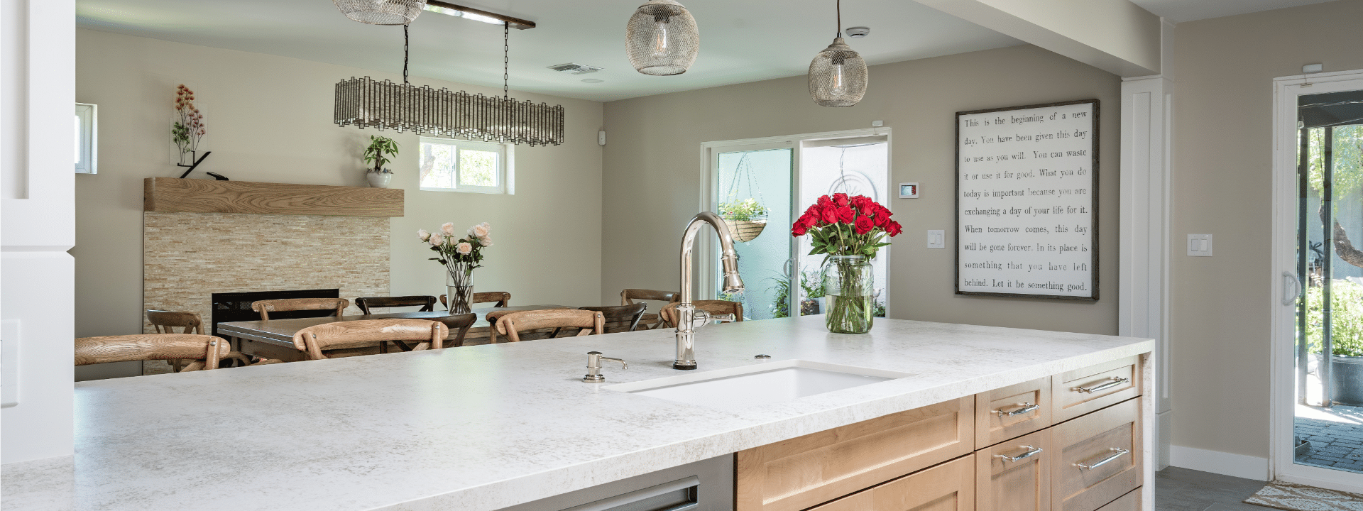 A modern kitchen in Paradise Valley, AZ, features a large white island with a built-in sink, light wood cabinets, and pendant lights. Ideal for home remodeling inspiration, it also includes a dining table, fireplace, wall sign, and red roses on the counter.