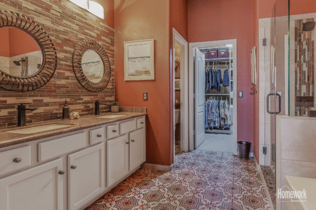Experience a stunning bathroom refresh with double sinks, round mirrors, patterned tile flooring, a mosaic backsplash, and white cabinets. Warm lighting and earthy tones flow into the walk-in closet visible through the open door.