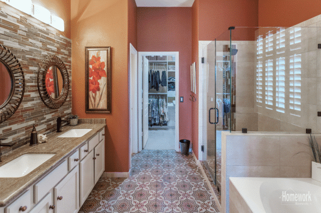 Spacious bathroom refresh with dual sinks, decorative mirrors, tiled backsplash, glass shower, bathtub, patterned floor tiles, and an open doorway leading to a walk-in closet with hanging clothes.