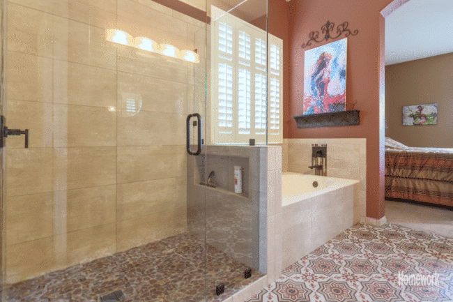 Modern bathroom refresh with a glass-enclosed shower featuring a pebble floor, a built-in tub with art above it, patterned tile flooring, and a view into a bedroom with a striped bedspread.