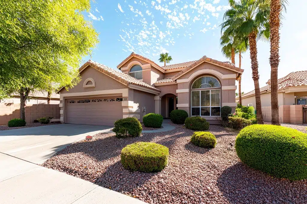 Single-story stucco home in Tempe, AZ with tan exterior, arched windows, and a two-car garage. The landscaped front yard features rocks, trimmed bushes, and palm trees beneath a bright, partly cloudy sky.