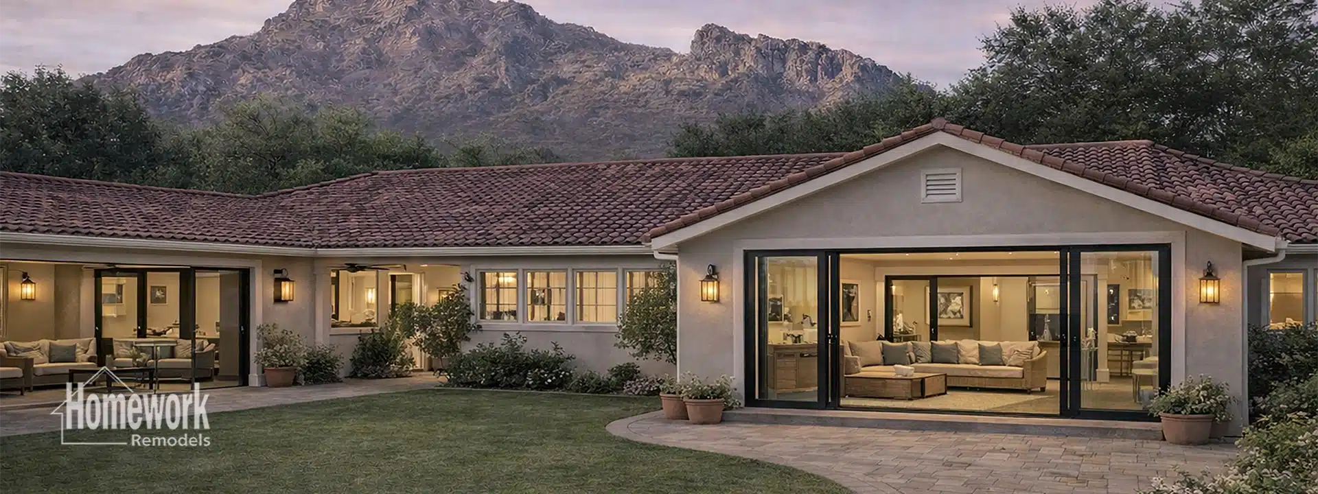 A single-story Arcadia house with large windows and sliding glass doors, surrounded by greenery and mountains. The warmly lit interior features a cozy living room. Logo reads Homework Remodels.