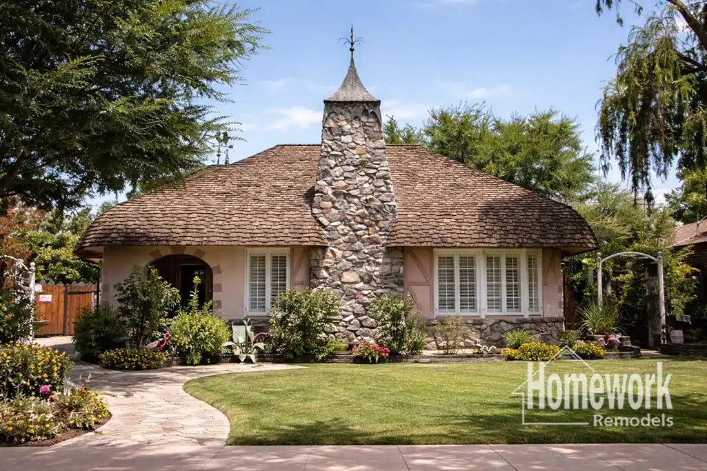 Charming cottage-style F.Q. house with a tall stone chimney, wood-shingle roof, and manicured lawn, surrounded by lush trees and colorful flowerbeds. Features clever historic home storage—a signature of Homework Remodels—logo in the bottom right corner.