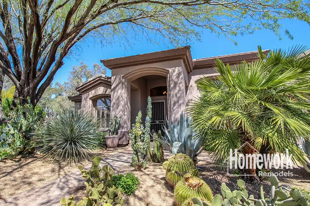 Single-story house with stucco exterior and arched entrance, surrounded by desert landscaping with various cacti and succulents in North Glendale. Blue sky above, and “Homework Remodels” logo in the bottom right corner—a showcase of home renovation.
