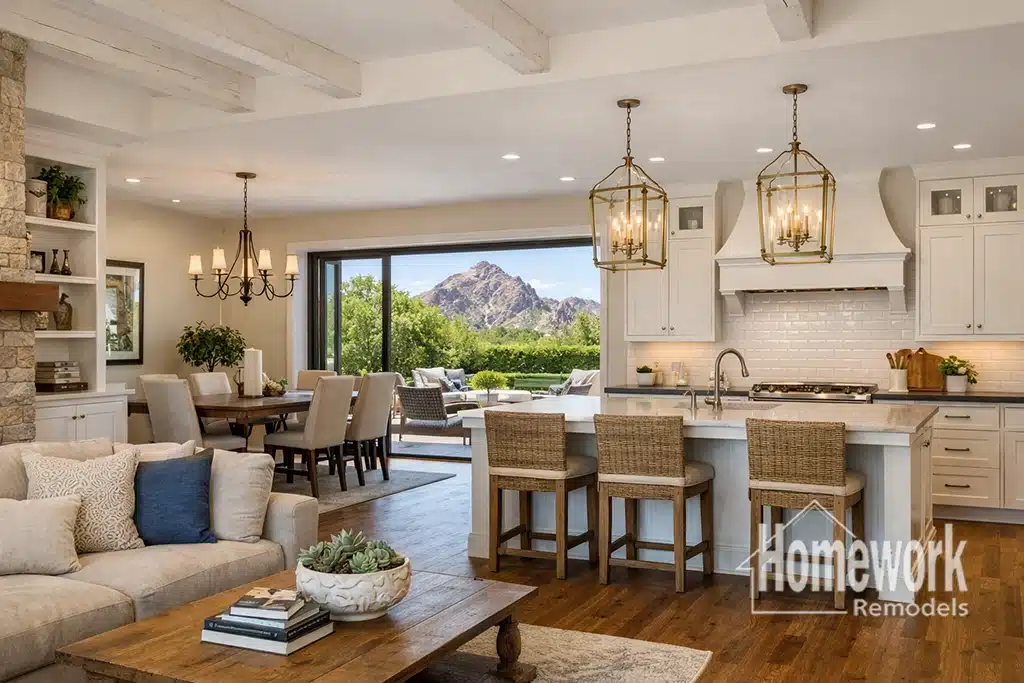 Open-concept living, Arcadia dining, and kitchen area with a stylish kitchen layout, wicker barstools, modern chandeliers, and large glass doors opening to a patio with mountain views. Homework Remodels logo in corner.