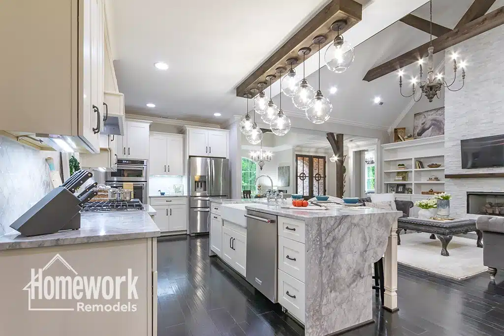A modern, open-concept Biltmore kitchen and living room with white cabinets, marble island, dark wood floors, industrial light fixtures, and a built-in fireplace. The “Homework Remodels” logo appears in the lower left corner.