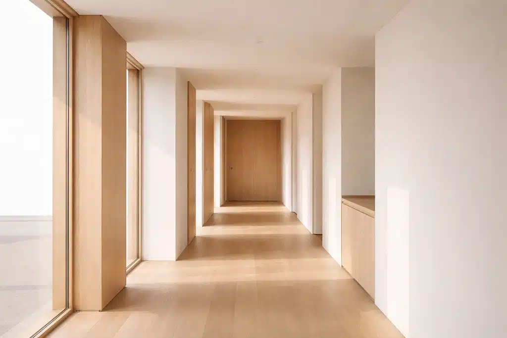 A minimalist hallway with light wood floors, large floor-to-ceiling windows on the left, white walls, and wooden doors highlights the importance of scope in design—sunlight streams in, creating a warm and airy atmosphere.