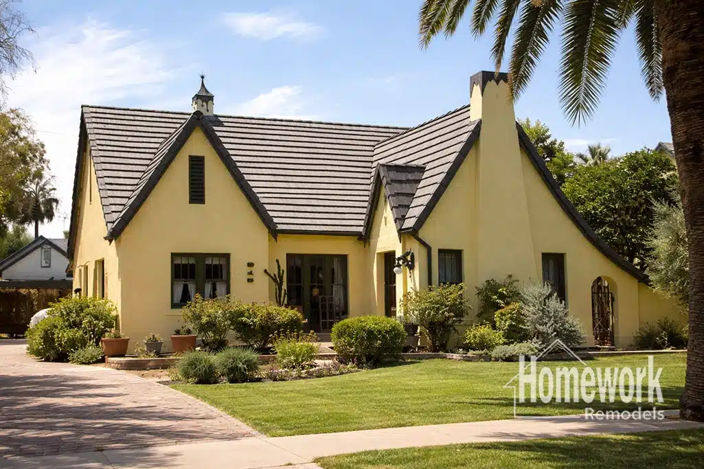 Charming yellow stucco house with steep, pointed rooflines in the Willo Historic District, featuring a green lawn, lush bushes, and palm tree. Sidewalk and Homework Remodels logo complete this showcase of expert remodeling.