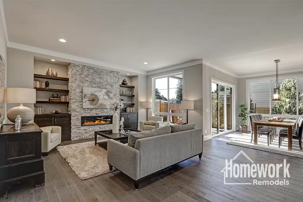 Modern living room with gray sofas, a stone fireplace, built-in shelves, and large windows. Perfect for Scottsdale homeowners who love open-concept floor plans. Adjacent dining area with sliding glass doors opens to a fenced backyard. “Homework Remodels” logo in corner.