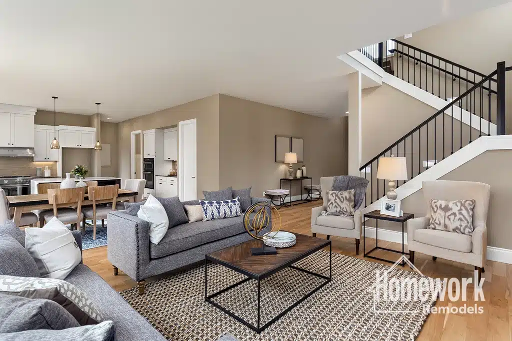 Scottsdale: Modern open-concept living and dining area with neutral tones, gray sofas, patterned pillows, wooden coffee table, light chairs, kitchen in background, and a staircase with black railings. Homework Remodels logo in corner.