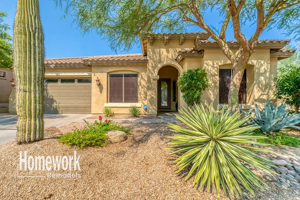 Phoenix Remodels: A beige stucco house with a two-car garage, arched entryway, and desert landscaping including cacti, agave, and gravel. The Homework Remodels logo is in the lower-left corner.