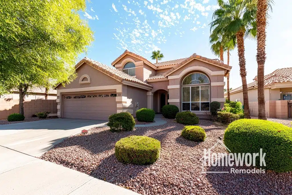 Tempe Homes: A stucco, two-story suburban house with a tile roof, arched windows, and a garage. The front yard features desert landscaping with rocks, trimmed bushes, and palm trees—showcasing smart home improvement to avoid common home mistakes. Homework Remodels logo is in the bottom right corner.