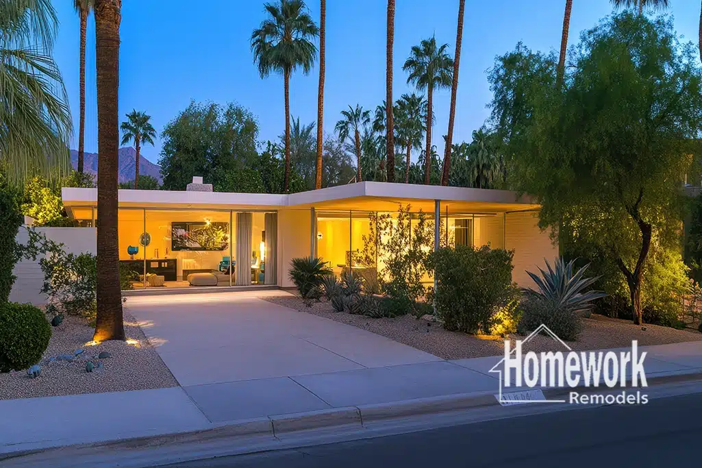 A modern, single-story Mid-Century Modern home with large windows glows warmly at dusk, surrounded by palm trees and desert landscaping. The Homework Remodels logo appears in the bottom right corner.