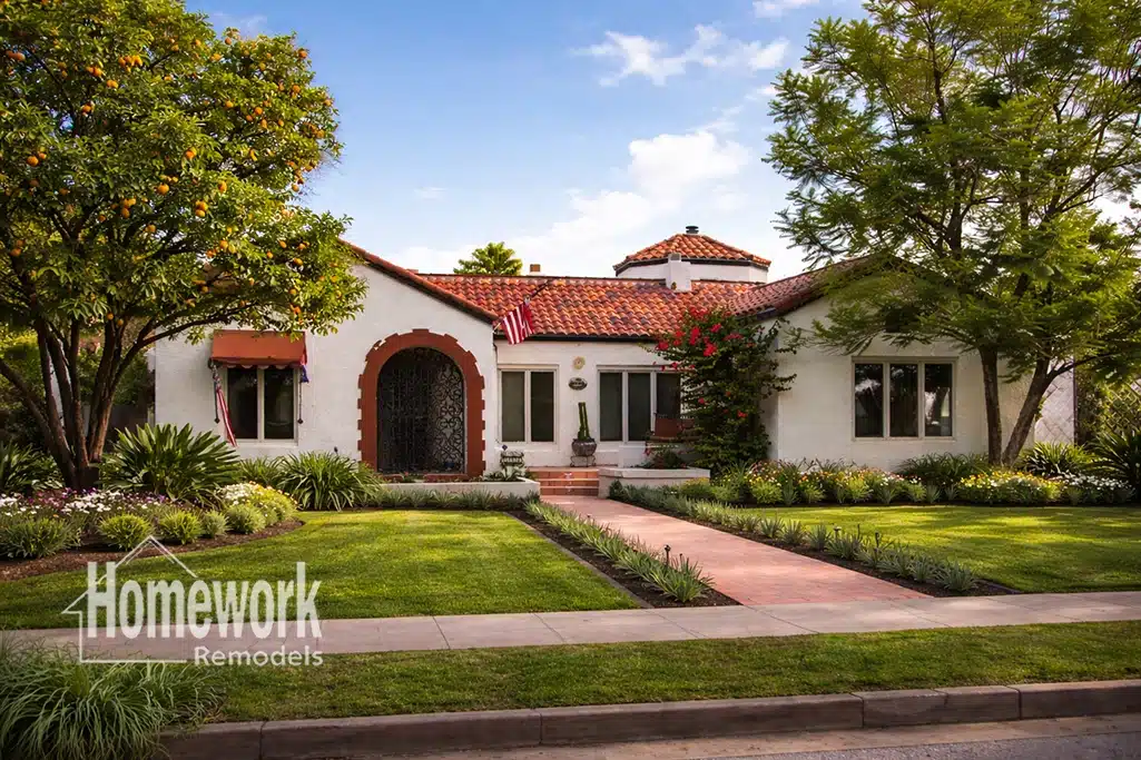 Whole-Home Remodeling in Roosevelt Historic District: A charming white stucco house with a red tile roof, arched entryway, manicured lawn, lush landscaping, and an American flag. Homework Remodels logo appears in the lower left corner.