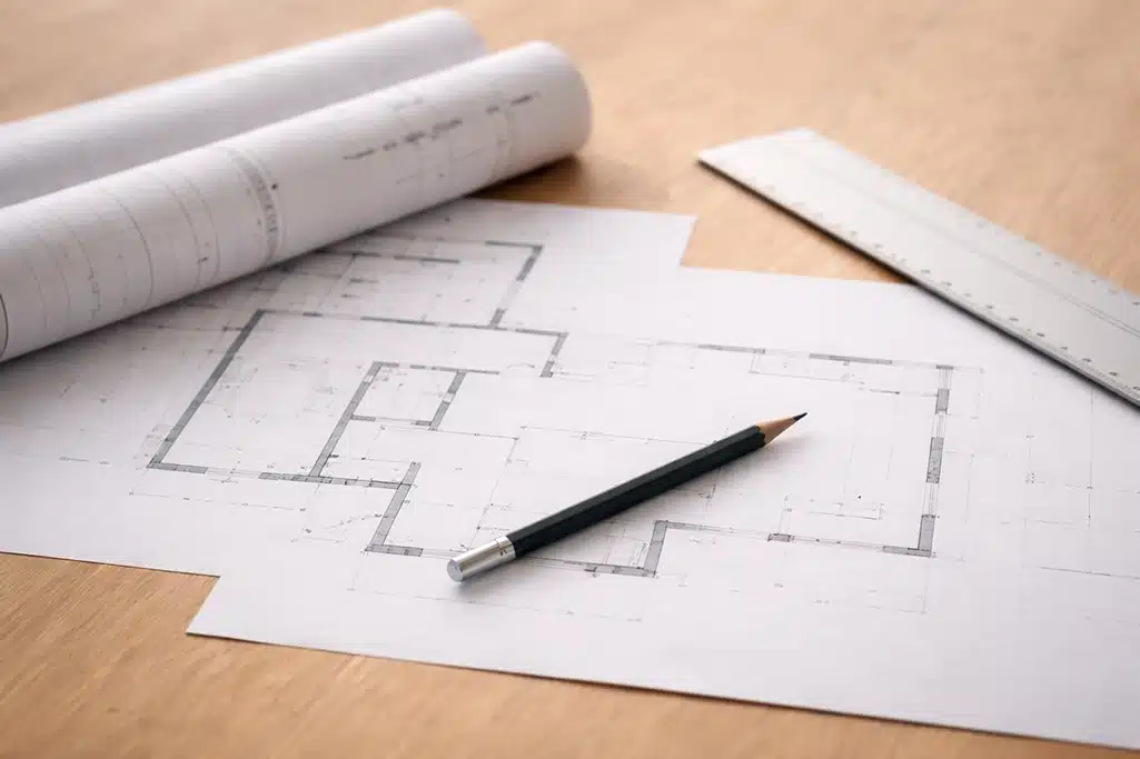 A pencil rests on an architectural floor plan drawing on a wooden table, with a rolled-up blueprint and a metal ruler nearby.
