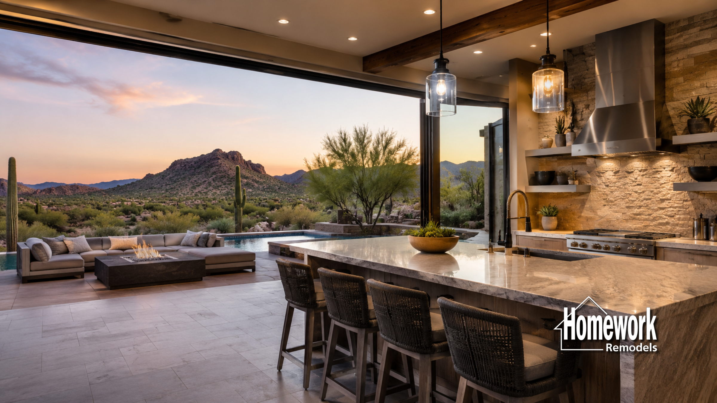 Luxurious open-concept kitchen and living space with stone accents overlooks a pool, patio, and scenic desert mountains at sunset. Homework Remodels logo is in the bottom right corner.