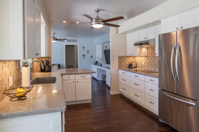 Modern kitchen with white cabinets, stainless steel appliances, granite countertops, tile backsplash, wood flooring, and ceiling fans. A bowl of fruit is on the counter, and crosses are displayed on a wall in the background.
