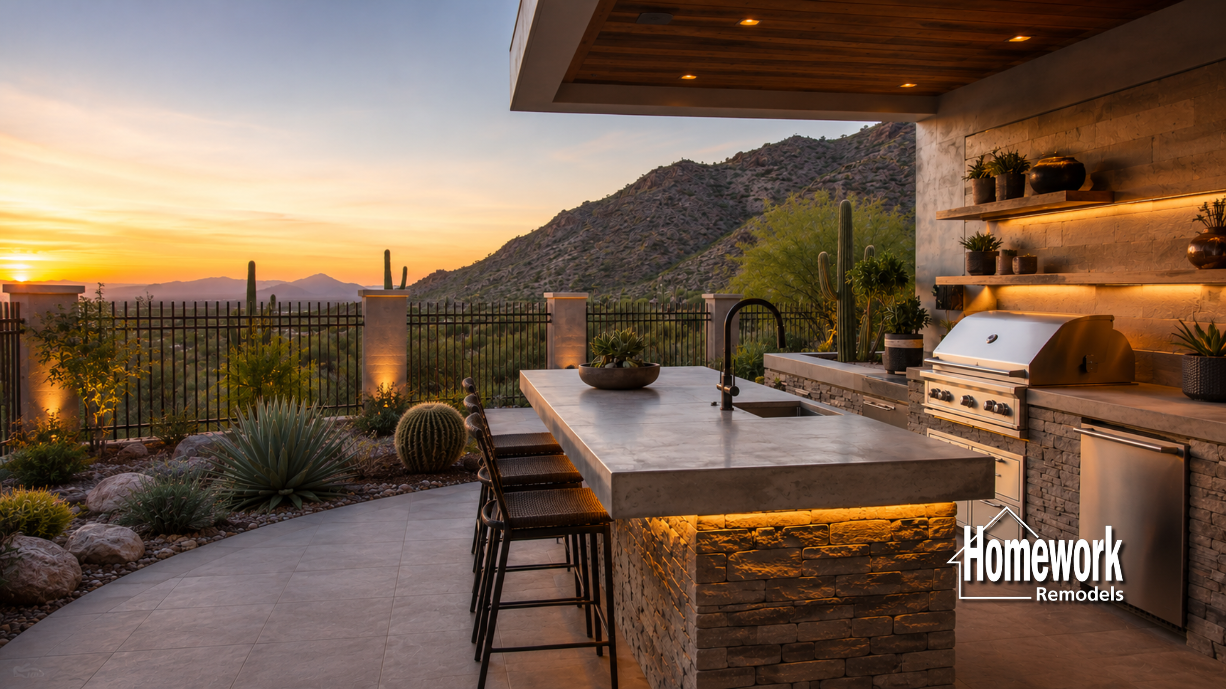 Modern outdoor kitchen design with stone counters, barstools, grill, and shelves set against a scenic mountain view at sunset. Warm lighting enhances this stylish patio—perfect for desert living. Homework Remodels logo in the bottom right corner.