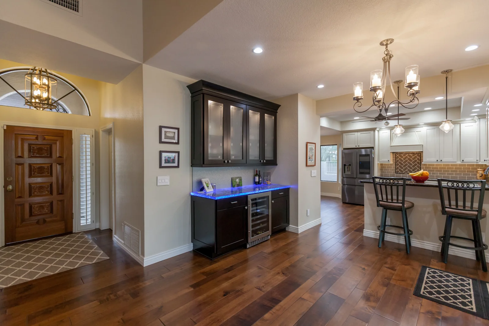 Custom dry bar with dark cabinetry, illuminated glass-front cabinets, a wine refrigerator, and a blue backlit glass countertop