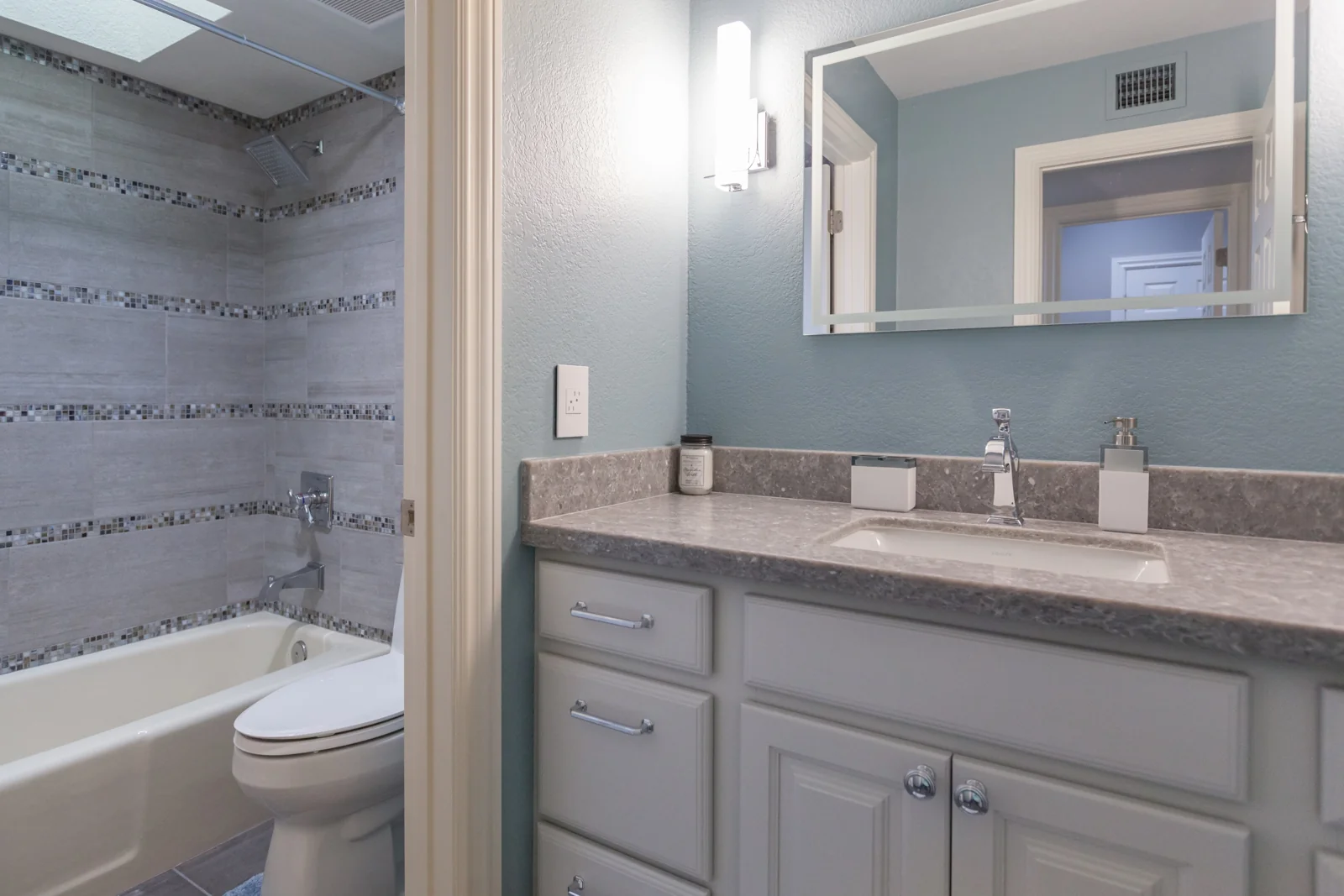 Updated guest bathroom with gray cabinetry, a framed mirror, new sconces, and patterned wall tile beside the retained bathtub