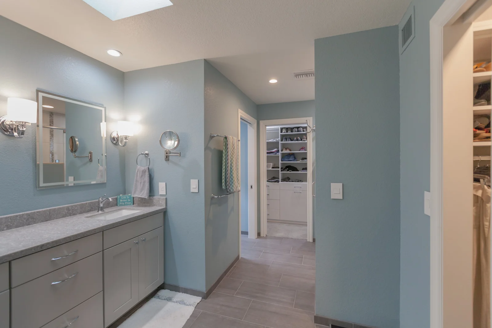 Remodeled primary bathroom with soft gray cabinets, quartz counters, blue walls, and an open route toward the closet