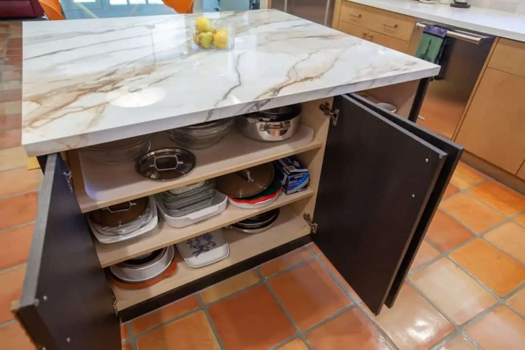 Open dark kitchen island cabinet with pull-out shelves storing bowls and dishes below a veined porcelain countertop