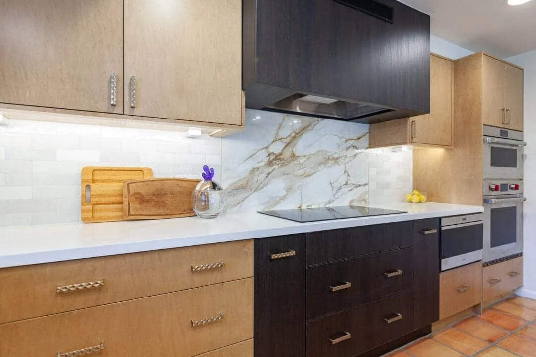 Remodeled cooking wall with a smooth-surface cooktop, dark hood and base cabinets, natural wood cabinetry, ovens, and veined porcelain backsplash